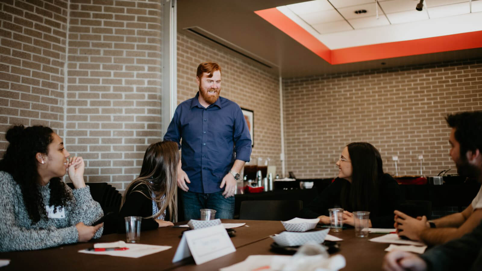 Professor, standing, interacts with students at a table.