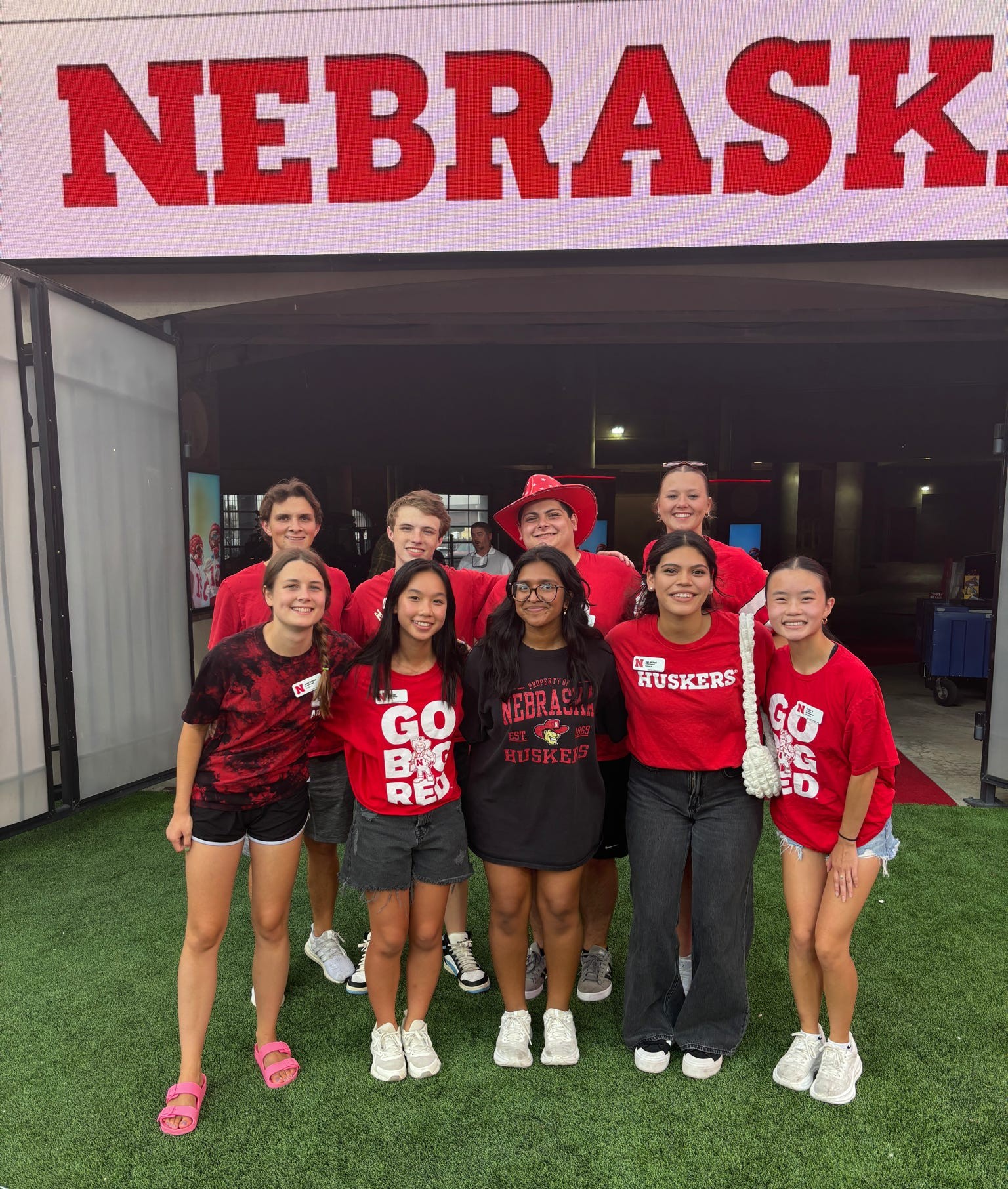 Group standing on grass, wearing red Nebraska Husker shirts, smiling in front of Nebraska sign.