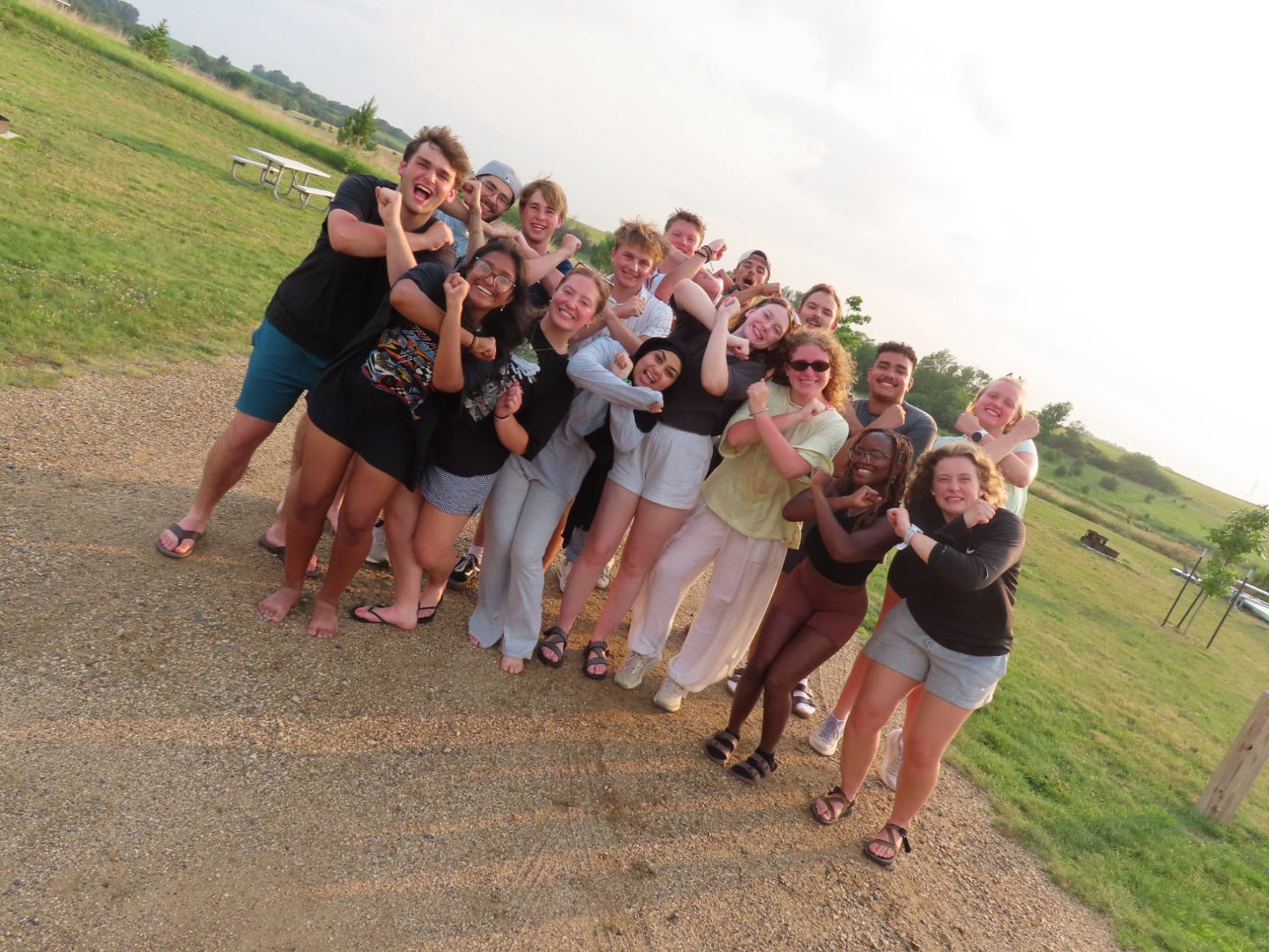 Nick with a group of people smiling and posing outdoors on grassy field, crossing arms in an X shape.