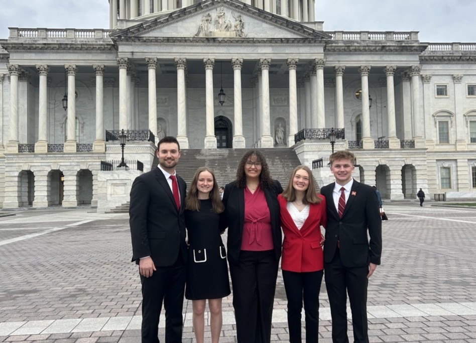 Group of five people in formal attire standing in front of a large government building.
