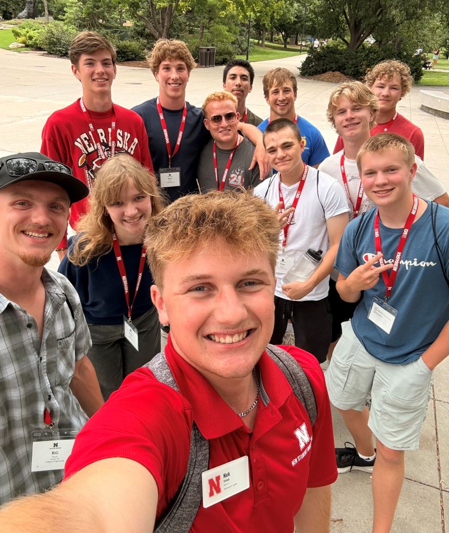 Group of young adults smiling outdoors, wearing name tags and lanyards, vibrant greenery in the background.