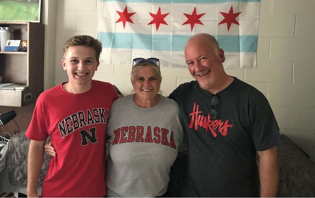 Three people smiling together, wearing Nebraska shirts, with a Chicago flag on the wall behind them.