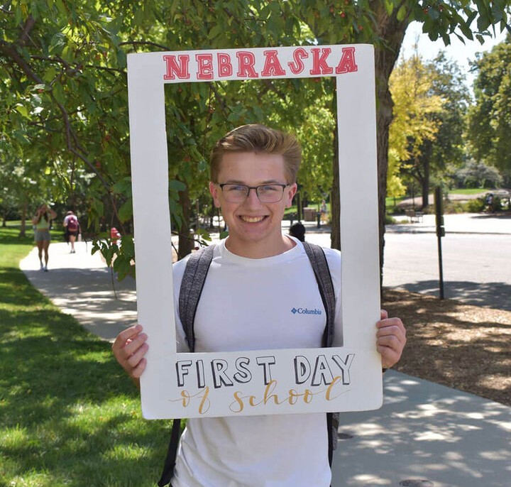 Nick holding a "Nebraska First Day of School" frame, smiling near trees and pathways.