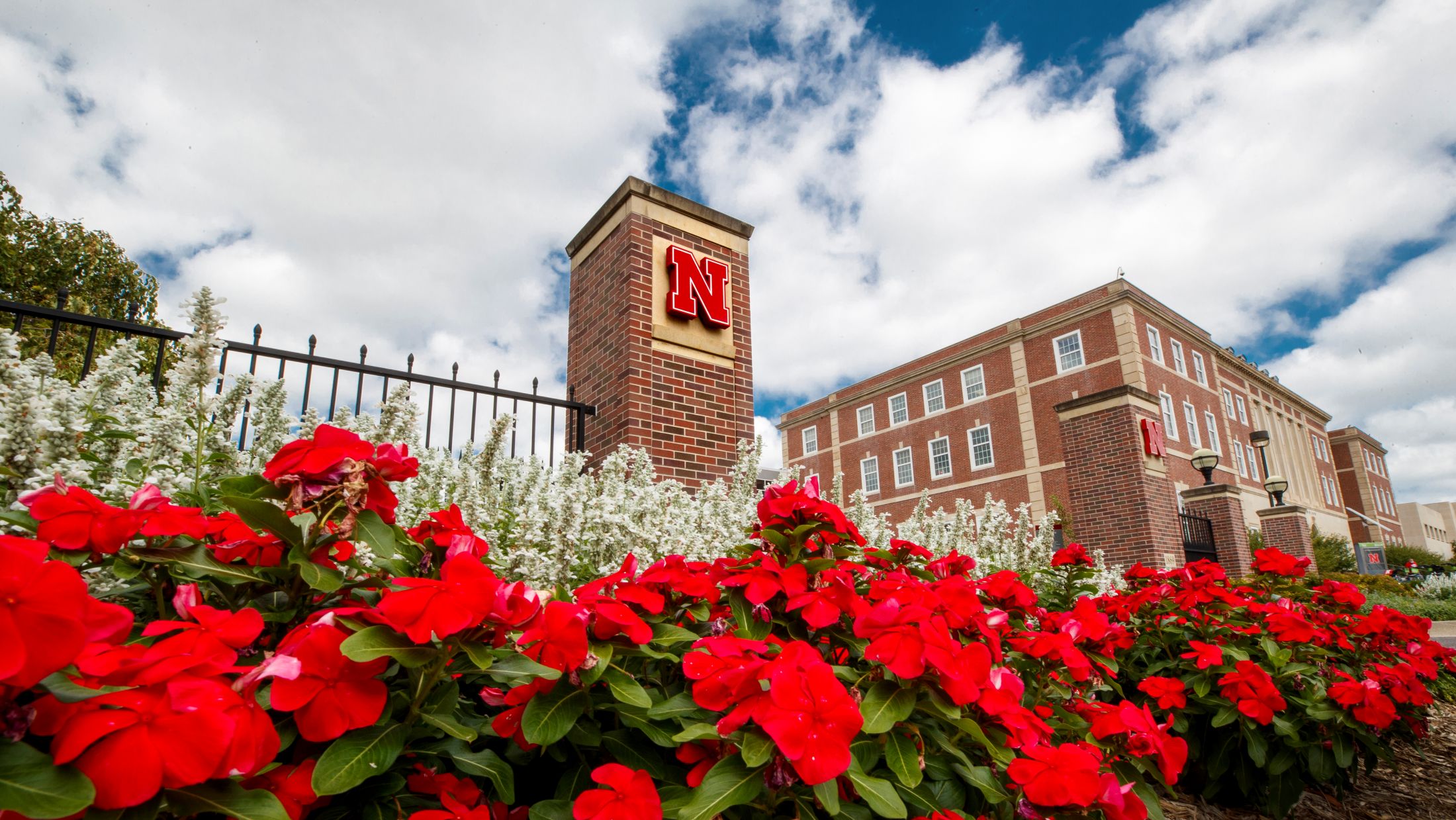Brick building with red flowers and pillar featuring a red "N" under a cloudy blue sky.