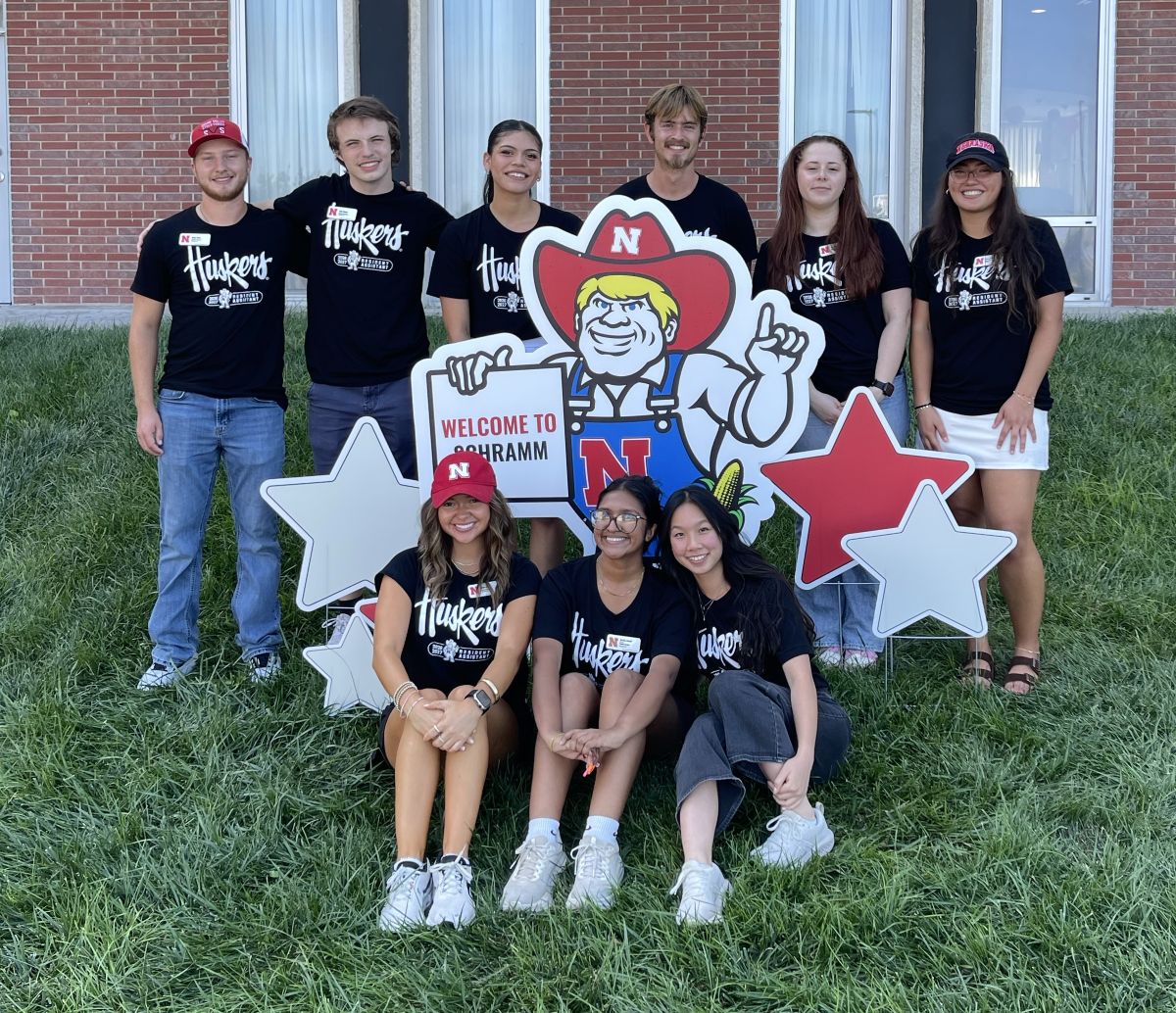 Group of people smiling, wearing Huskers shirts, posing outdoors with a Nebraska mascot sign and stars.