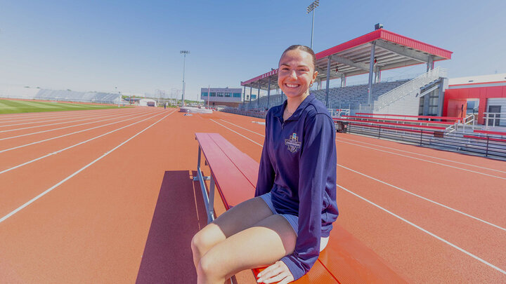 Berlyn Schutz, a white woman with brown hair sits in a blue track and field sweater and shorts, on a red bench on a red track.